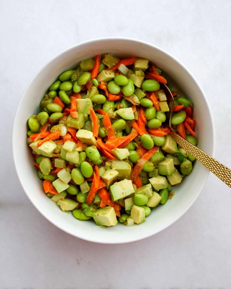 A white bowl filled with a mix of three layers of diced vegetables and beans: bright green edamame beans, small cubes of pale green avocado, and thin strips of orange carrots, all mixed together showing varied rough and smooth textures; a gold textured spoon is placed inside the bowl, resting diagonally from the right side. The bowl sits on a white marbled surface. Photo taken with an iphone --ar 4:5 --v 7