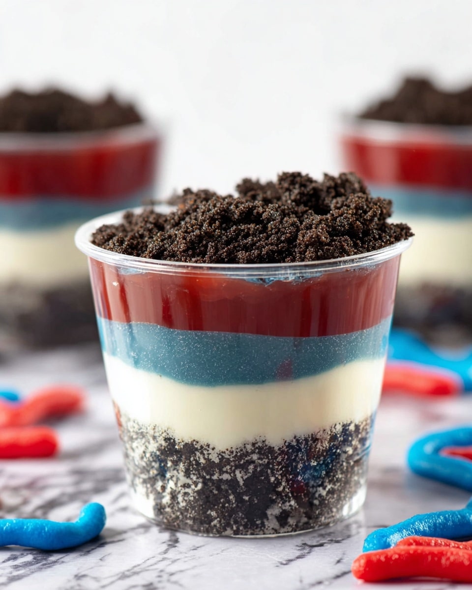 A clear cup shows a five-layer dessert starting with a crumbly dark cookie base at the bottom, followed by a smooth, thick blue layer that is glossy. Above this is another crumbly dark cookie layer, topped by a creamy white layer with a smooth texture. Another crumbly cookie layer sits on top of the white cream, followed by a shiny red gel-like layer, and finally a thick, crumbly dark cookie topping with rough texture at the very top. The dessert is placed on a white marbled surface with a few red and blue candy worms around it, and there are similar cups blurred in the background. Photo taken with an iphone --ar 4:5 --v 7