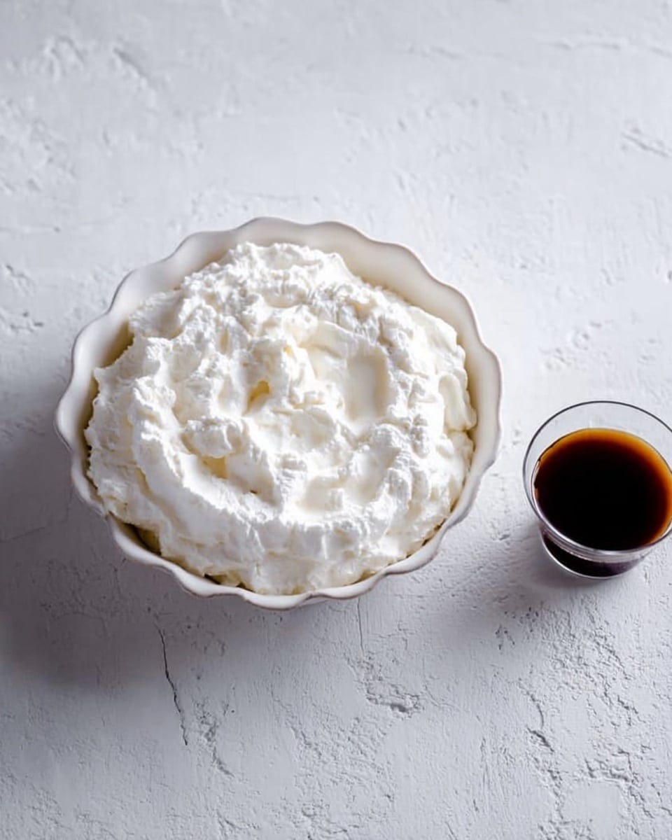A white bowl with a scalloped edge filled with thick, fluffy white whipped cream showing soft swirls and texture on top, placed on a white marbled surface. Next to the bowl, on the right side, is a small clear glass cup filled with dark brown coffee. The scene is bright and simple with soft natural light. Photo taken with an iphone --ar 4:5 --v 7