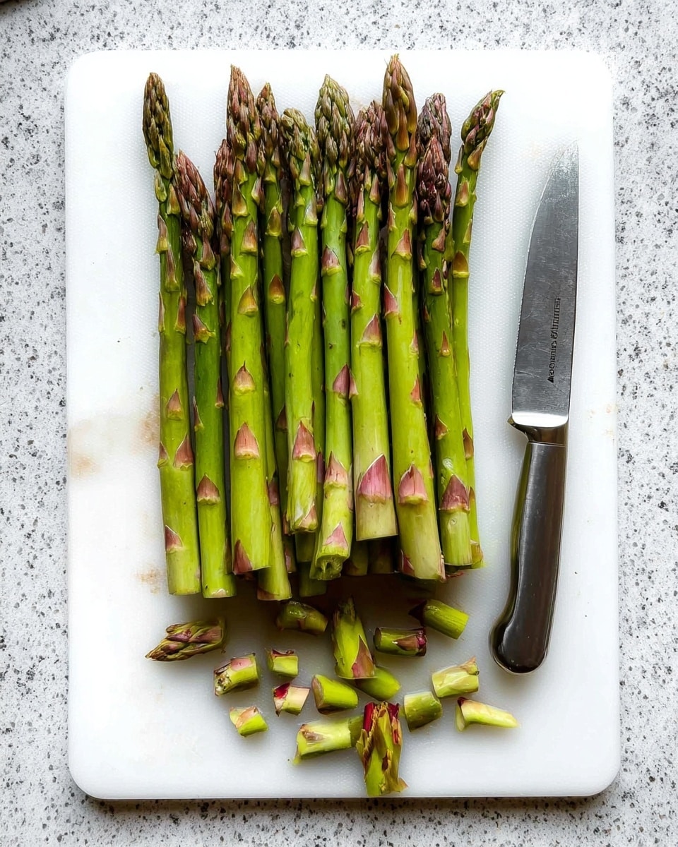 A bunch of green asparagus spears lined up vertically on a white cutting board, with the bottom tough ends already removed and small pieces scattered at the base. The asparagus tips are darker green with purplish hues, and the stems show some brownish patches. To the right side of the cutting board, there is a silver knife with a sharp blade resting flat. The cutting board is placed on a white marbled textured surface. photo taken with an iphone --ar 4:5 --v 7