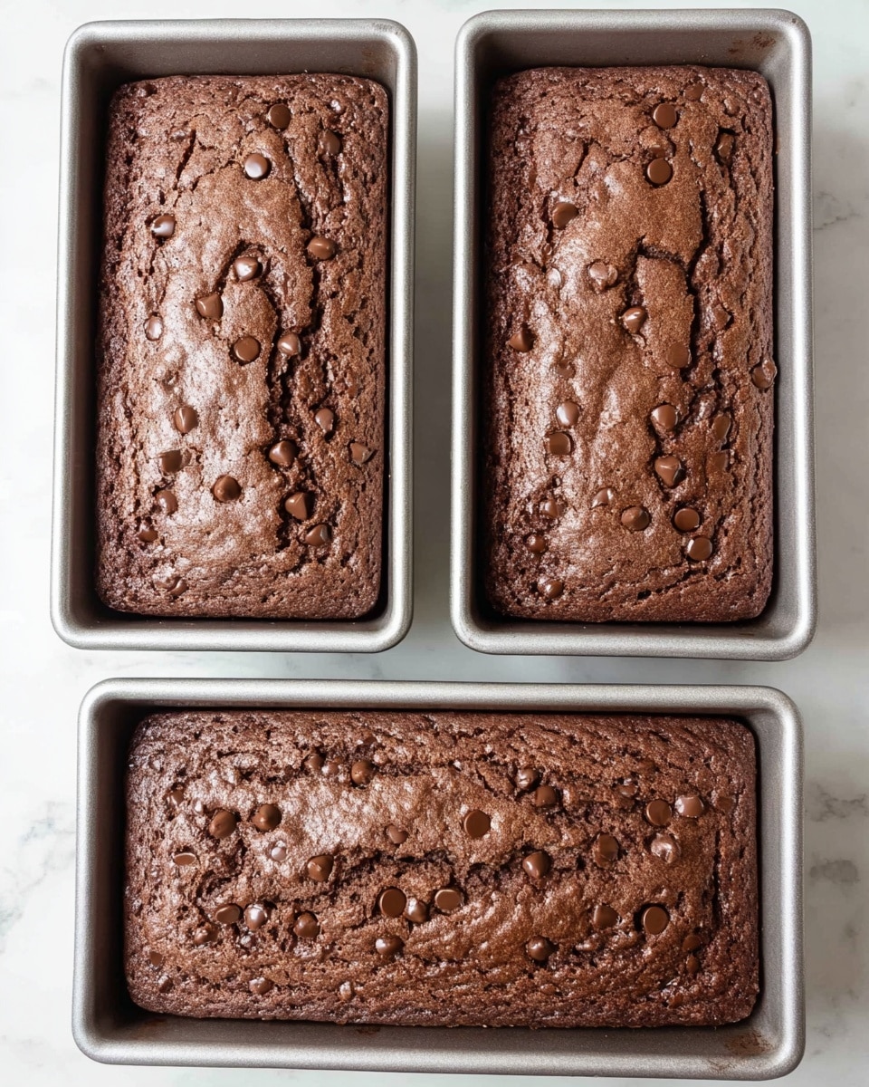 Two rectangular chocolate cakes rest side by side on a white marbled surface, each in a light gray metal baking pan. The cakes have a rich brown color with a slightly cracked but soft texture on top, dotted with numerous melted chocolate chips scattered evenly across the surface. The edges of the cakes are slightly darker, showing they are well baked. The background is clean and simple, emphasizing the moist and inviting look of the chocolate cakes. photo taken with an iphone --ar 4:5 --v 7