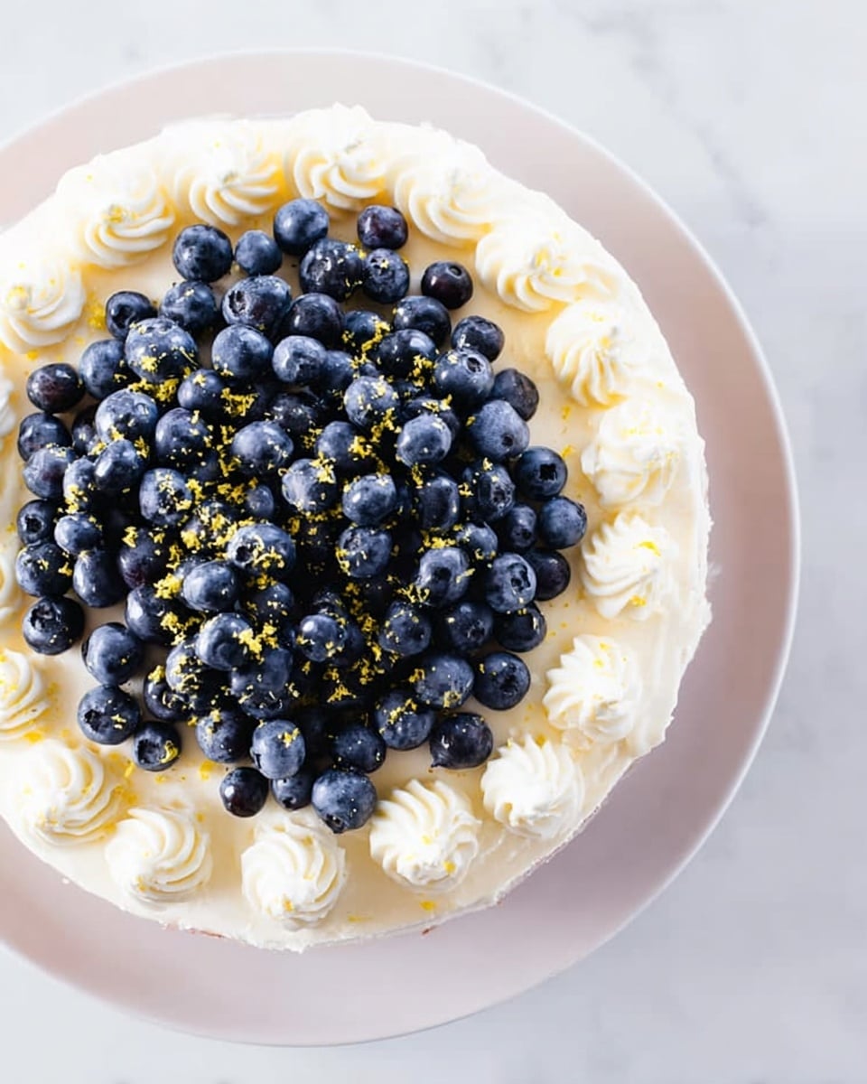 A round cake placed on a white plate with a white marbled surface underneath. The cake has a thick layer of white cream frosting covering the top. Around the edge, there are evenly spaced swirls of the same white cream. The center of the cake is covered with a generous pile of fresh blueberries scattered closely together. Small yellow zest pieces are sprinkled lightly over the blueberries, adding color contrast. The image has bright, natural lighting. photo taken with an iphone --ar 4:5 --v 7