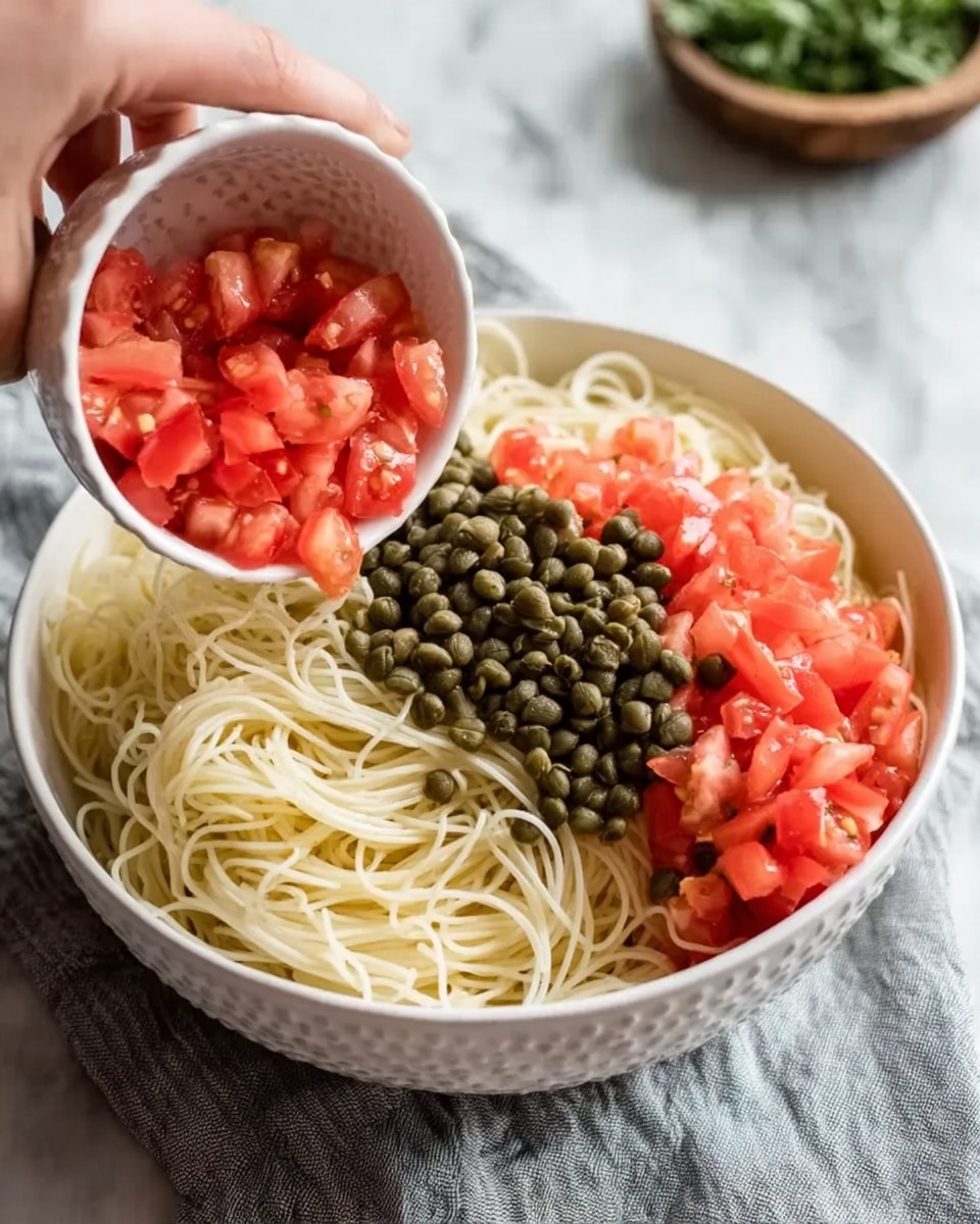 A white bowl filled with thin cooked noodles is placed on a white marbled surface with a light gray cloth underneath. On one side of the noodles, there is a pile of small green capers, and on the other side, fresh chopped red tomatoes are being poured from a white textured small bowl held by a woman's hand. Some green herbs are blurred in the background. photo taken with an iphone --ar 4:5 --v 7