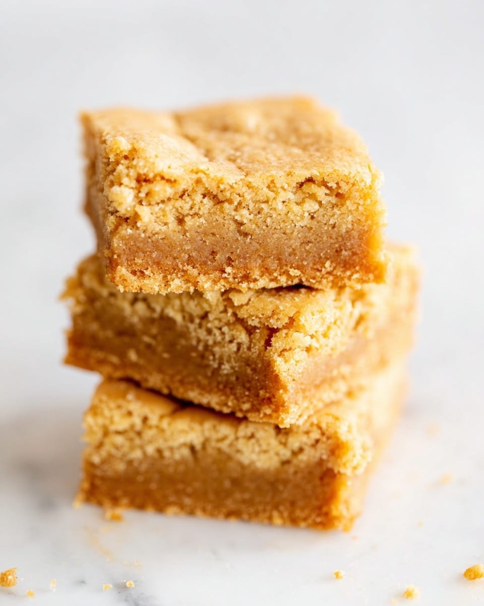 A close-up view of a single-layer square baked item with a light golden brown top and a slightly darker brown edge, showing a moist and dense texture inside. The cut-out corner reveals the inside, which is crumbly but soft, resting on a white marbled surface with a few crumbs scattered near the base. Photo taken with an iphone --ar 4:5 --v 7