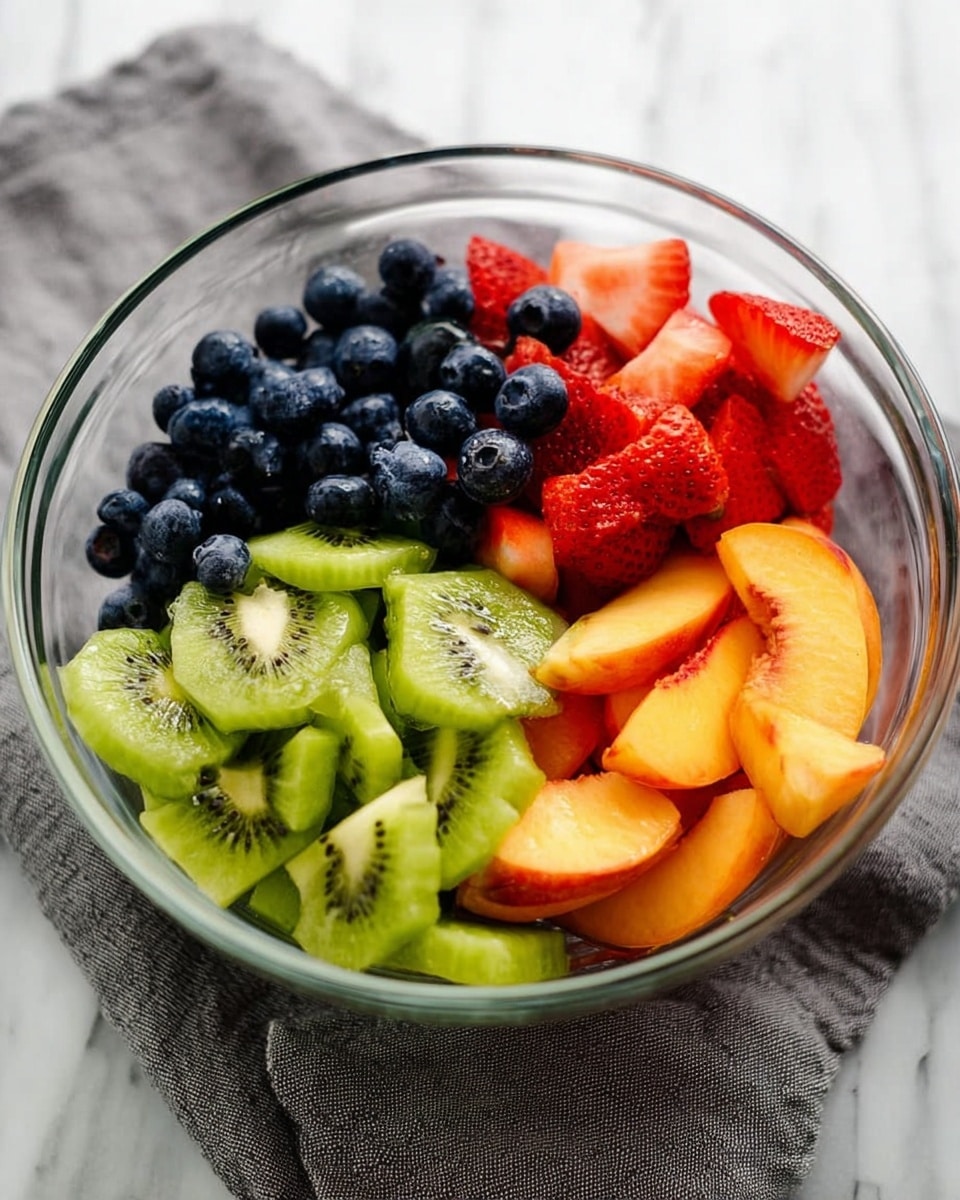 A clear glass bowl sits on a gray cloth over a white marbled surface, filled with four separate piles of fresh fruit. On the left, there are small round blueberries with a deep blue color. Below them, bright green kiwi slices, cut in half and arranged in layers showing their soft, slightly translucent texture and tiny black seeds. To the right of the kiwis, red strawberries are sliced and stacked, showing the juicy inside and seeds on the outside. On the top right, orange peach slices curve neatly in a small stack. The colors of the fruits are vibrant, and their textures vary from smooth to slightly fuzzy. photo taken with an iphone --ar 4:5 --v 7