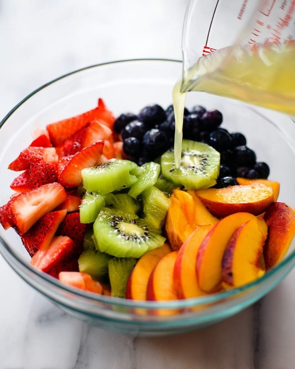 A clear glass bowl filled with five types of fresh fruit layered around the inside. At the bottom left are bright red sliced strawberries. Next to them on the bottom right are orange and yellow mixed peach slices. Above the peach slices on the right are larger orange peach wedges. At the top center are small dark blue blueberries. In the middle left are light green kiwi slices with a soft, juicy texture. A light yellow liquid is being poured over the fruit from a measuring cup above. The bowl sits on a white marbled surface. Photo taken with an iphone --ar 4:5 --v 7