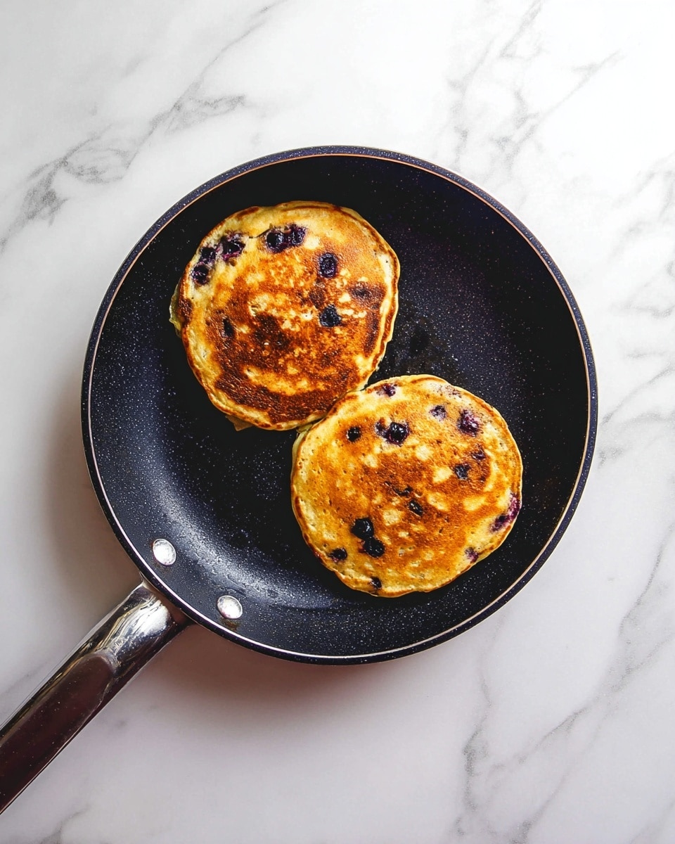 Two golden-brown pancakes with dark purple spots from blueberries are inside a black frying pan with a dark handle. The pancakes show a slightly uneven cooked texture with some light and dark areas. The pan sits on a white marbled surface. photo taken with an iphone --ar 4:5 --v 7