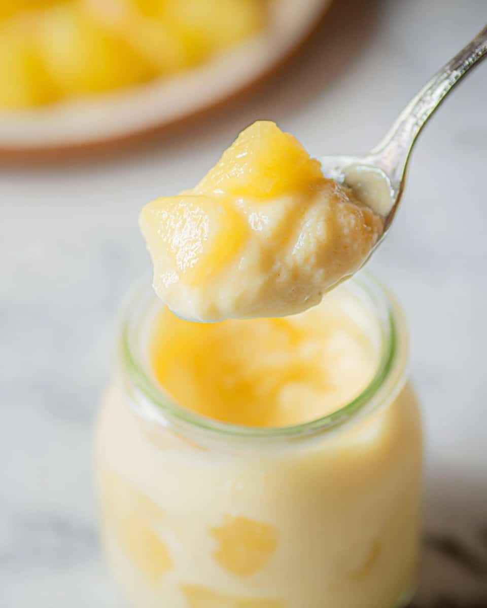 A close-up image shows a spoon holding a creamy light yellow dessert with small pieces of yellow fruit, lifted above a small clear glass jar filled with the same dessert and fruit mix. The dessert looks smooth and thick with a soft texture. The background has a white marbled texture with a blurred hint of more yellow fruit. Photo taken with an iphone --ar 4:5 --v 7