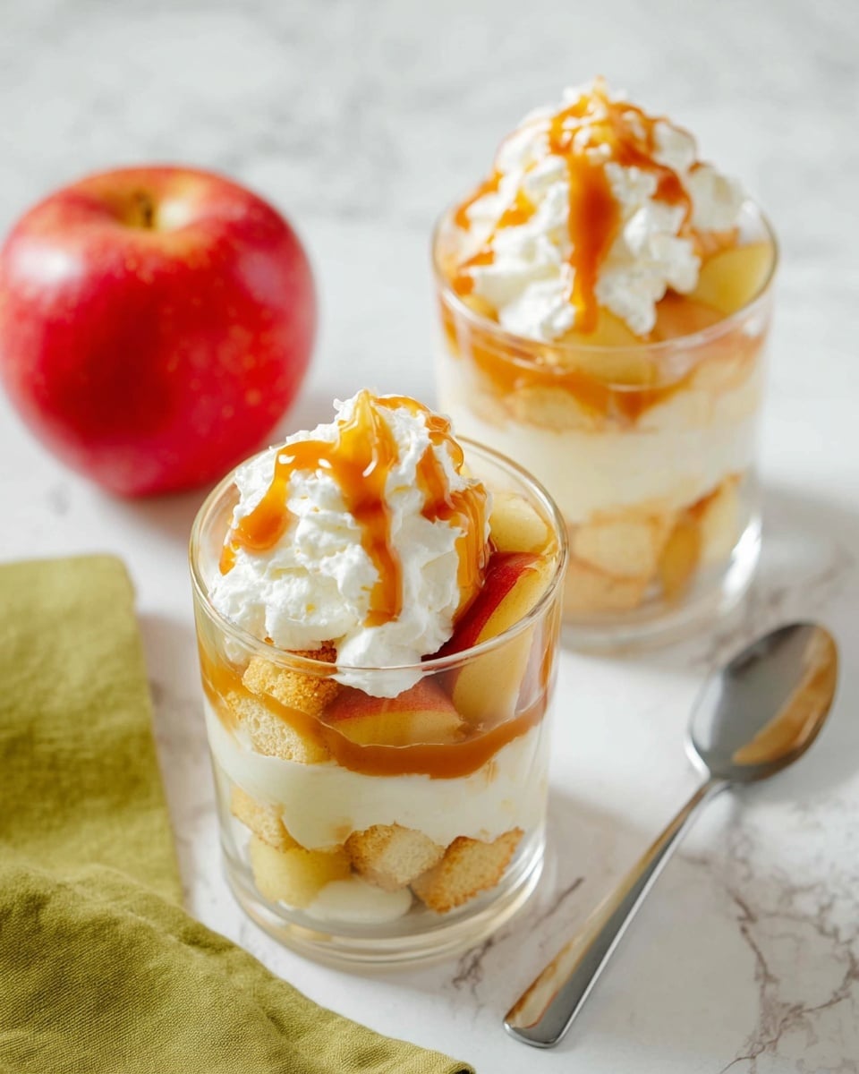 Two clear round glasses contain layered desserts placed on a white marbled surface. Each dessert shows three main layers: at the bottom, small golden cake cubes; in the middle, white creamy filling with some apple pieces; on top, a mound of white whipped cream drizzled with amber caramel sauce. In the background, there is a bright red apple on a green cloth, and a silver spoon lies beside the glasses. The overall color palette is warm with white and golden tones, and the scene has soft natural light. photo taken with an iphone --ar 4:5 --v 7