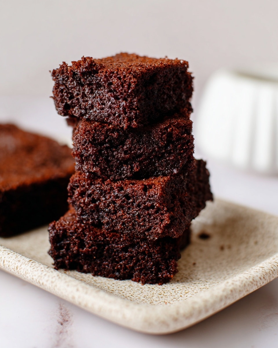 The image shows a small stack of four dark brown chocolate brownies with a soft, moist texture. The brownies have a rough, crumbly top and sides with tiny air holes visible. They are placed closely together on a speckled light beige rectangular plate which sits on a white marbled surface. The background is blurred but keeps a light, neutral tone. Photo taken with an iphone --ar 4:5 --v 7