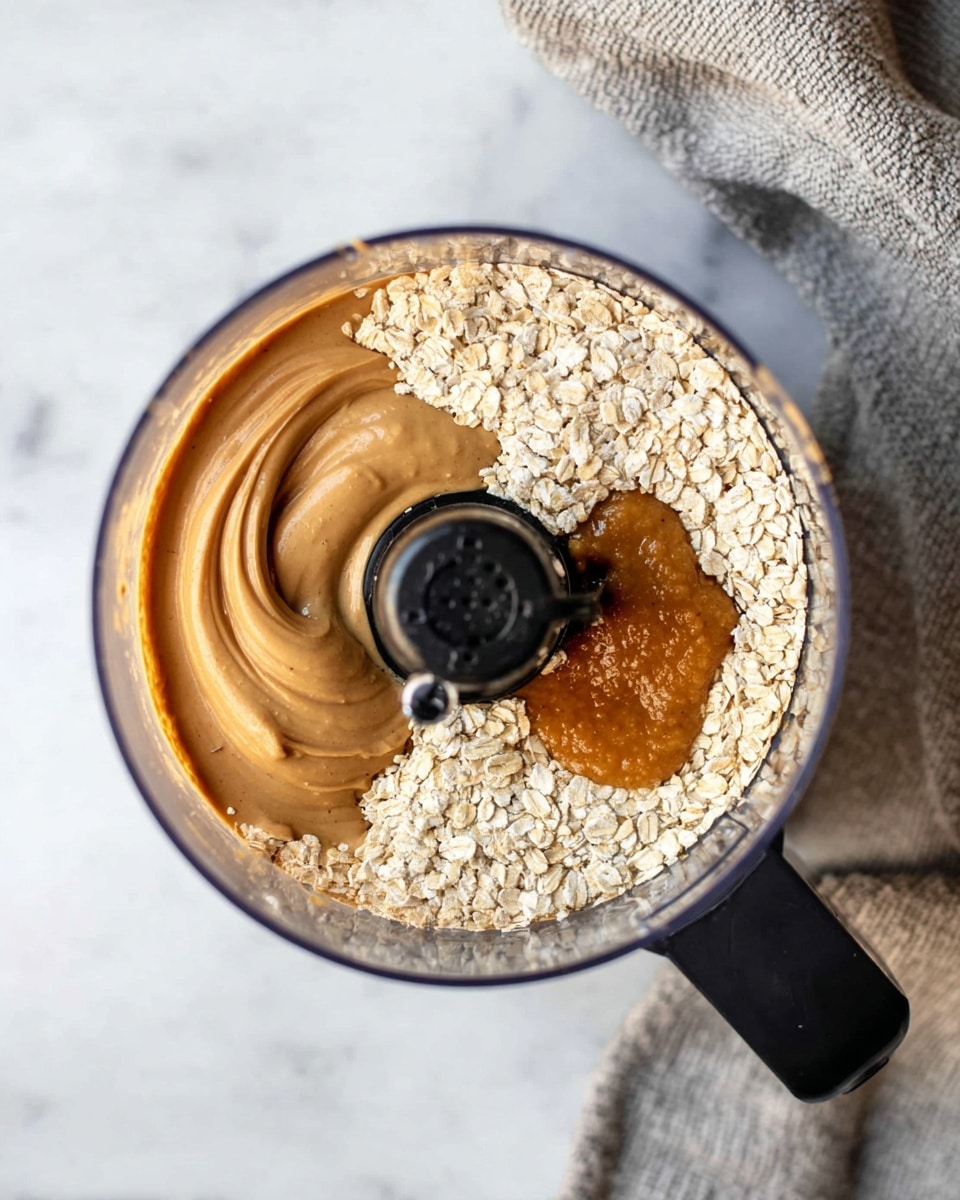 A top view of a clear food processor bowl sitting on a white marbled surface, showing three main layers inside: a smooth light brown peanut butter layer on the top right, a thick golden honey layer on the bottom right, and a dry, light beige oat layer covering the bottom left. The central black processor blade is visible, dividing the ingredients slightly. Part of a gray textured cloth is visible in the upper right background. Photo taken with an iphone --ar 4:5 --v 7