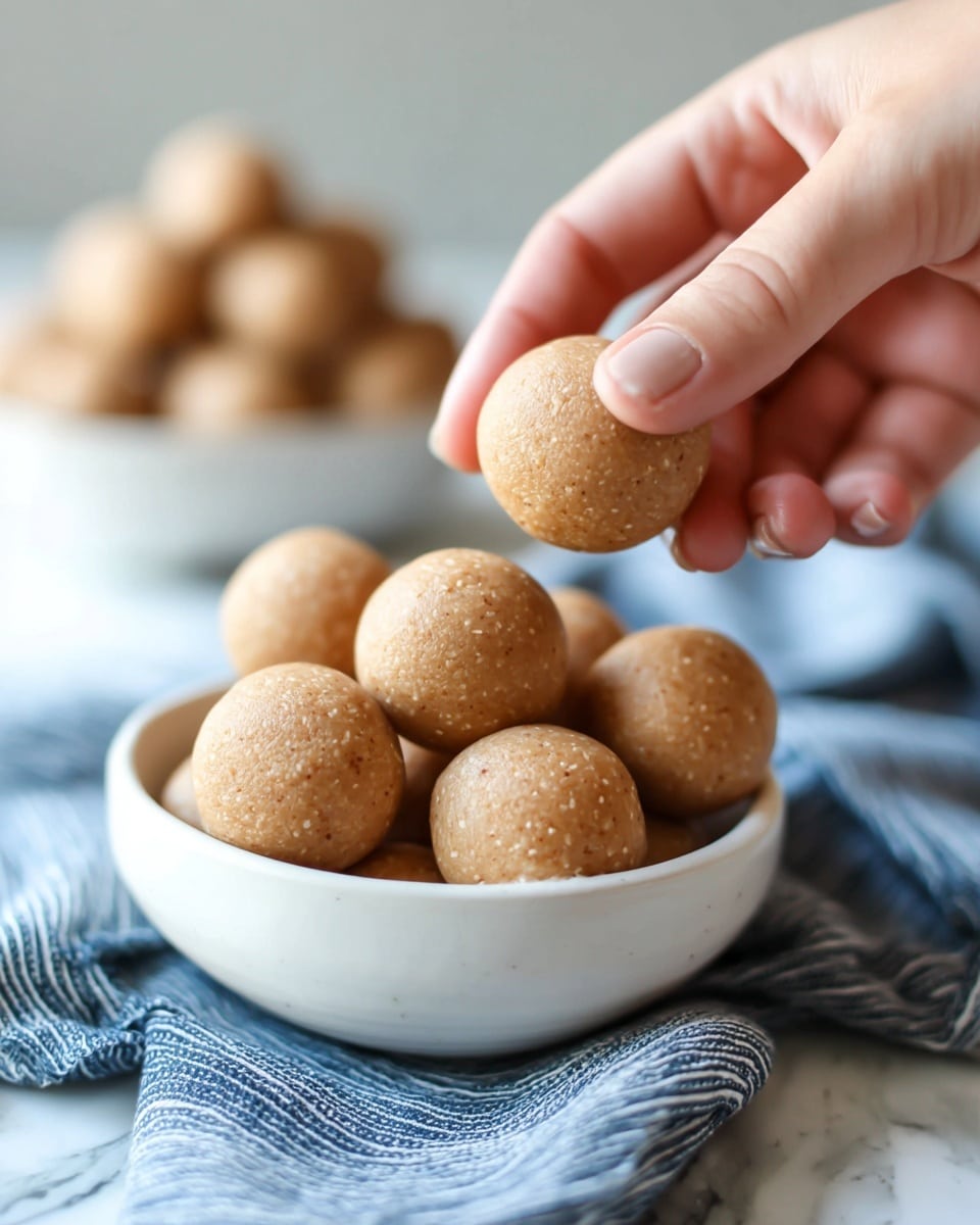 A close-up photo shows a white bowl full of smooth, light brown round balls with a slightly rough texture. A woman's hand is gently picking up one ball from the top of the bowl. In the blurry background, there is another white bowl with the same balls, placed on a white marbled surface. A blue and white striped cloth lies beneath the bowl in the foreground, adding a soft texture to the scene. The lighting is natural and soft, highlighting the details of the balls and the skin of the woman's hand. photo taken with an iphone --ar 4:5 --v 7