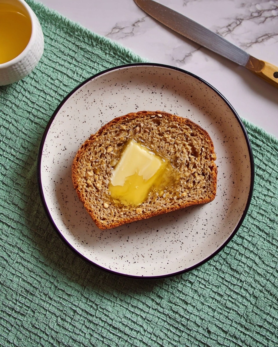 A single slice of brown bread with a rough, grainy texture is placed in the center of a white plate with small black specks and a thin black rim. The top layer of the bread is spread with melted yellow butter that looks soft and slightly shiny. The plate sits on a green towel with a waffle pattern, and next to it, there is a white marbled surface with a butter knife and a small white bowl containing more yellow butter. Photo taken with an iphone --ar 4:5 --v 7