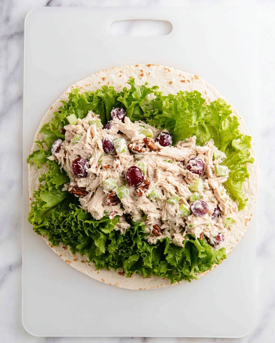 A soft, round tortilla lies flat on a white plastic board with a handle at the top. On the bottom half of the tortilla, there is a layer of fresh green leafy lettuce with ruffled edges, providing a crisp base. Above the lettuce, there is a thick spread of creamy chicken salad mixed with small pieces of chopped celery, halved red grapes, and bits of pecans, all coated in a light, white dressing. The texture on top looks chunky and creamy with bits of meat and fruit, creating a contrast with the smooth tortilla and leafy greens. The background is a white marbled surface. photo taken with an iphone --ar 4:5 --v 7