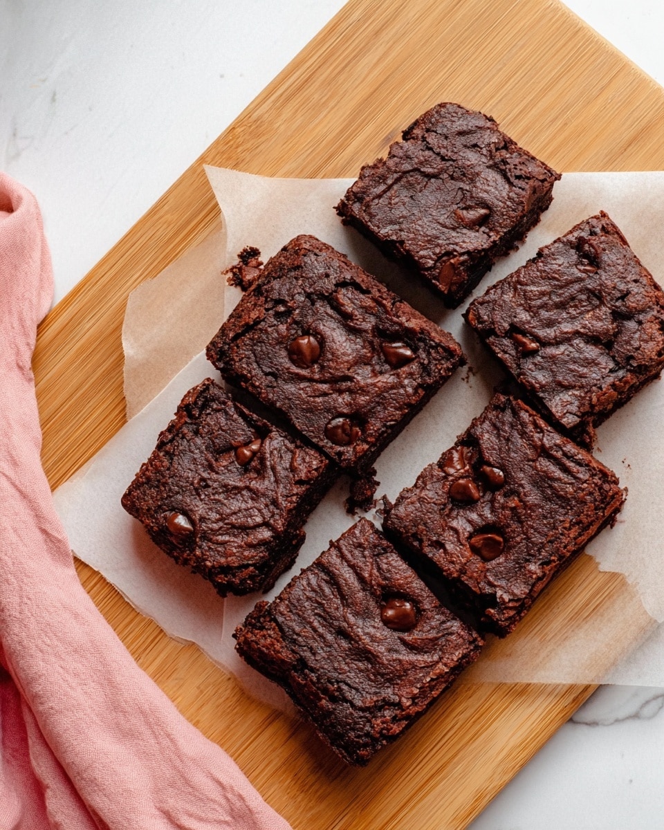 Six square pieces of dark brown chocolate brownies are placed on white parchment paper on a light wooden cutting board. The brownies have a slightly cracked, rough textured top with visible chocolate chips embedded in the surface. The edges of each brownie are slightly raised and the inside looks moist and dense. A corner of a pink cloth is visible in the bottom left corner on a white marbled background. photo taken with an iphone --ar 4:5 --v 7