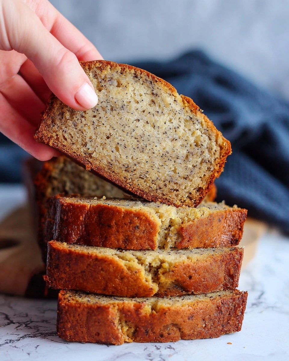 A close-up showing a woman's hand holding a slice of banana bread with a golden brown crust and soft, moist, light brown interior speckled with darker banana bits, above a stack of similar slices on a white surface with a white marbled texture background and a blurred dark blue cloth in the back, photo taken with an iphone --ar 4:5 --v 7