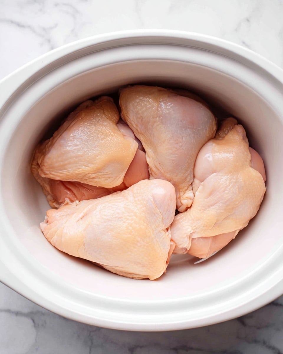 Inside a white slow cooker, there are four large raw chicken pieces placed closely together, filling most of the space. The chicken has a smooth, pale pink skin with slight shine and natural muscle lines visible. The slow cooker is set on a white marbled surface that adds a clean and bright background to the image. Photo taken with an iphone --ar 4:5 --v 7