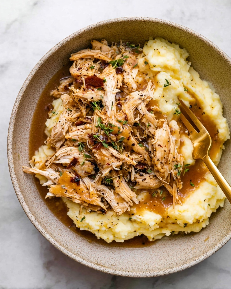 A bowl with two main layers: the bottom layer is creamy mashed potatoes that are light yellow and soft in texture, and on top is shredded cooked chicken with a light brown color, seasoned with herbs and black pepper, slightly shiny from a brown gravy sauce drizzled over it; small green herb pieces are scattered on the chicken, and there is a golden fork resting in the bowl on the right side, partially covered by the chicken. The bowl is placed on a white marbled surface. photo taken with an iphone --ar 4:5 --v 7