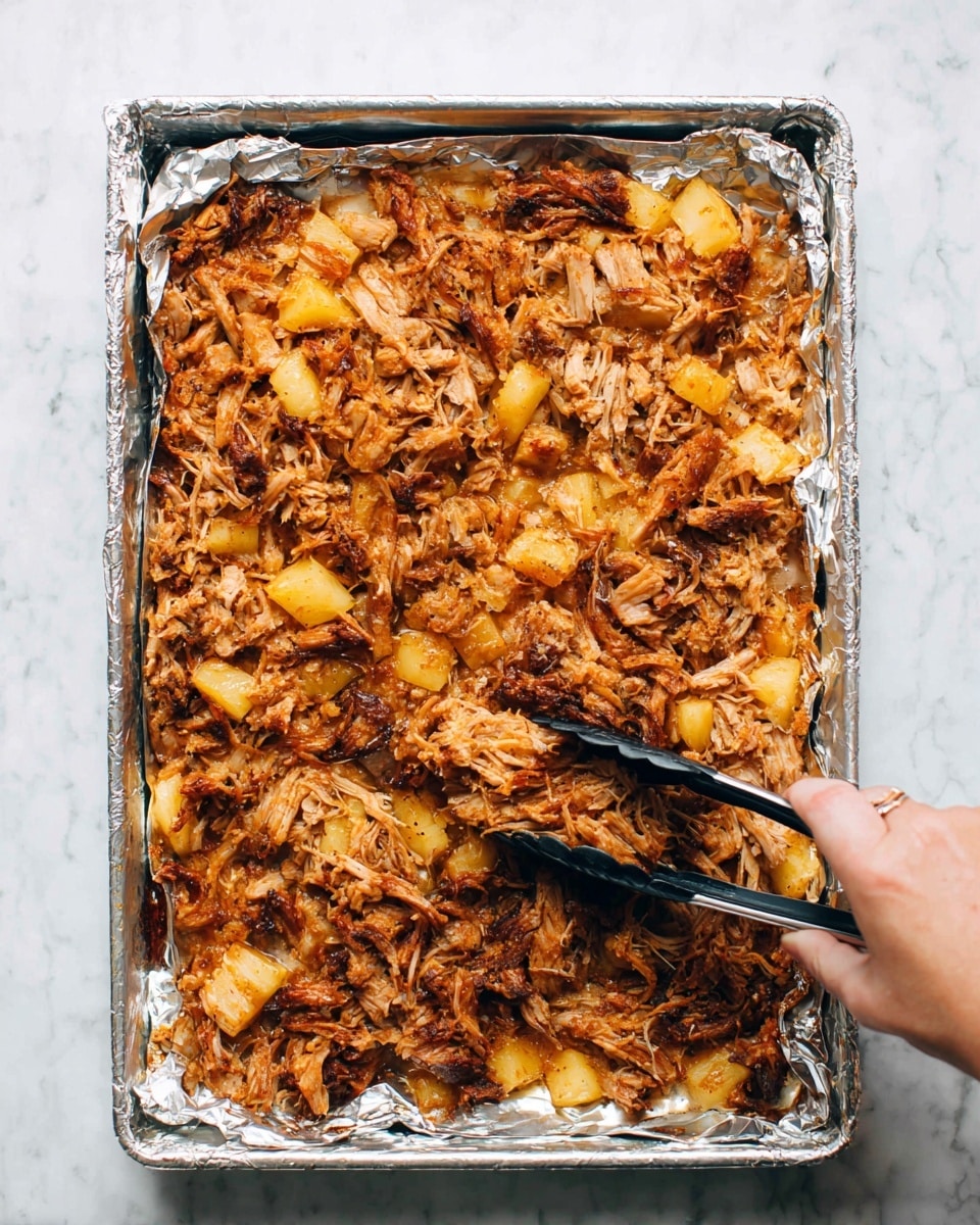 A rectangular baking tray lined with crinkled foil holds a single layer of shredded, browned cooked meat mixed with small chunks of softened yellow pineapple. The mix has spots of darker caramelized edges and lighter golden parts, creating a textured, uneven surface. A black and silver tong in a woman's hand is shown slightly lifting some of the meat near the bottom right corner. The tray sits on a white marbled surface. photo taken with an iphone --ar 4:5 --v 7