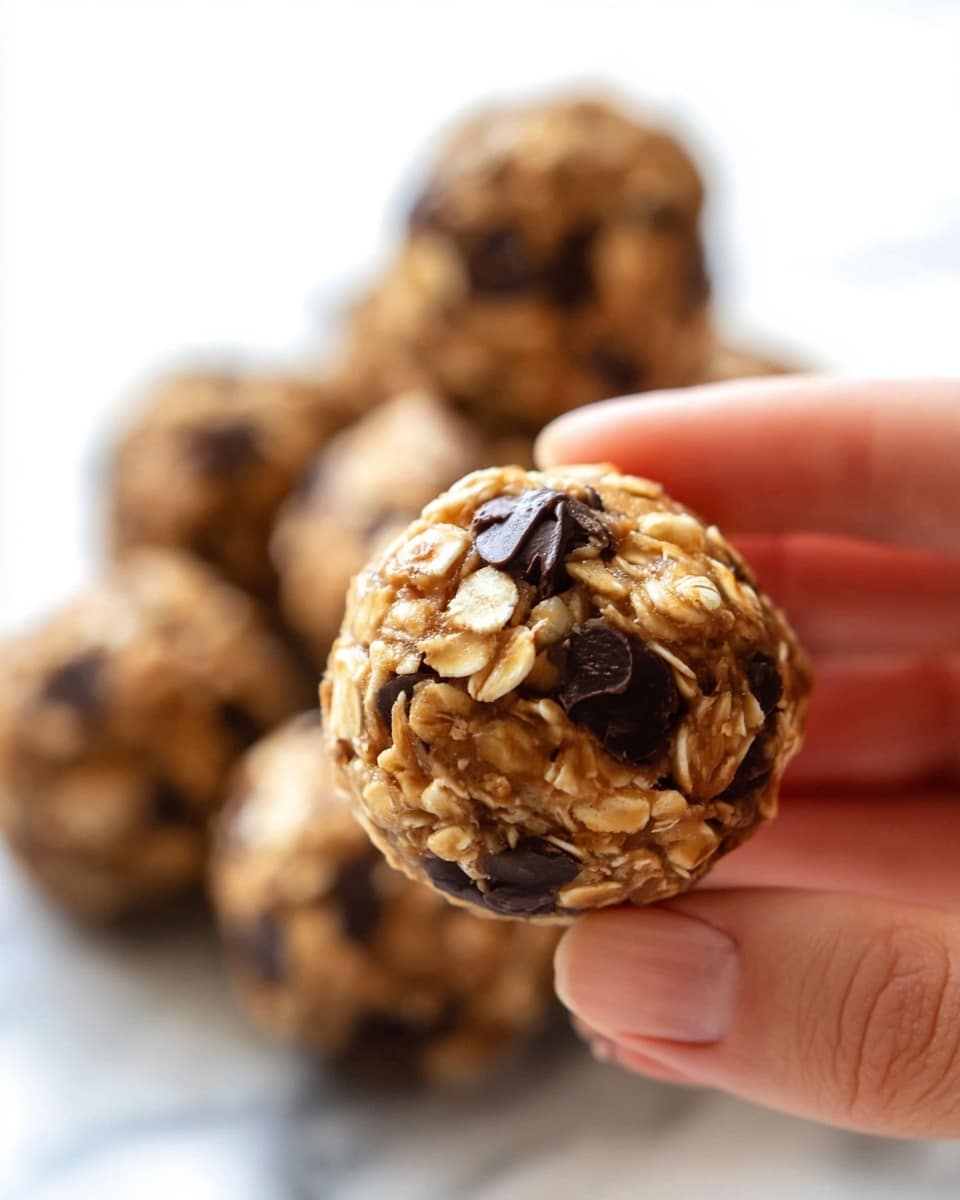 A close-up image shows a woman's hand holding a small round snack ball made of oats and chocolate chips. The ball has multiple layers of small oats, light brown sticky mixture, and dark chocolate chips scattered throughout its surface, giving a rough textured look with some shiny spots. In the background, there is a pile of similar snack balls softly blurred on a white marbled surface. Photo taken with an iphone --ar 4:5 --v 7