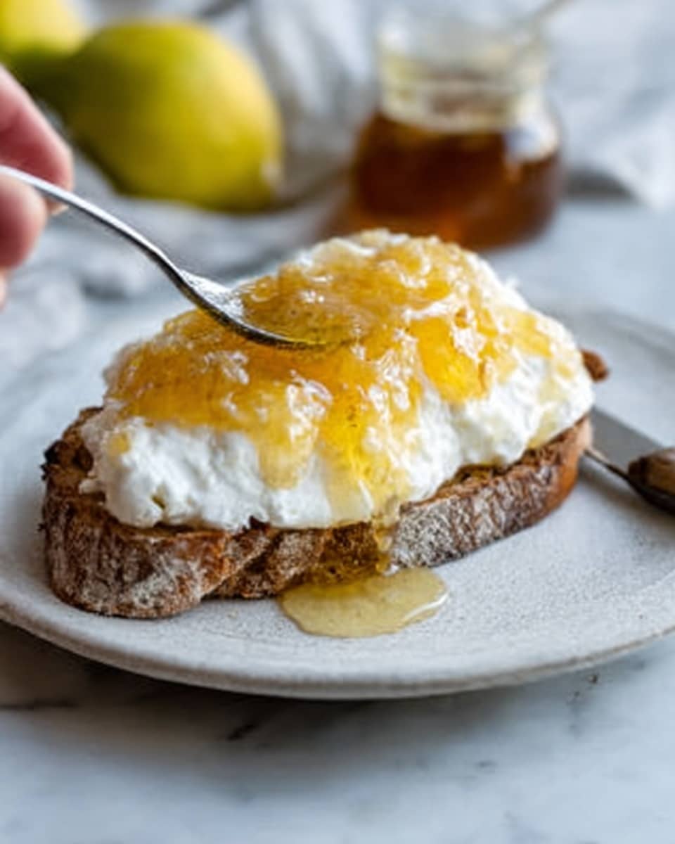 A close-up image shows a rustic piece of bread topped with a thick layer of white creamy cheese that looks soft and fluffy. On top of the cheese, there is a layer of translucent golden honey with visible small pieces giving it texture. Honey drips slightly off the side of the bread. The bread is placed on a clean white plate with a knife resting next to it, all set on a white marbled surface. A woman's hand is holding the bread from one side. photo taken with an iphone --ar 4:5 --v 7