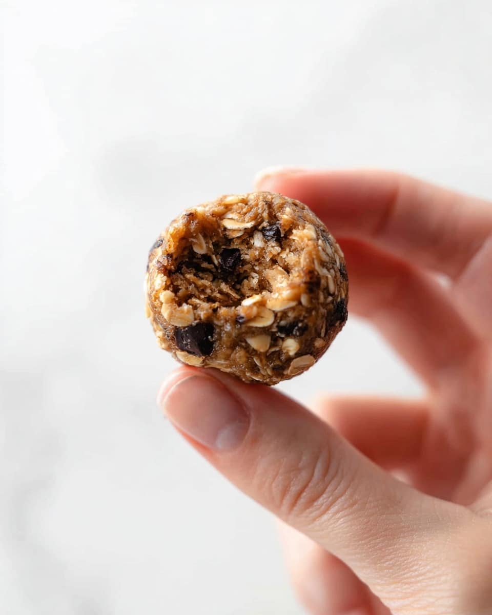 A woman’s hand holding a single small round energy bite with a rough texture, showing oats and small dark chocolate pieces embedded in the light brown, dense mixture. The bite has a visible bite mark on one side, revealing its soft inside. The background is a simple white marbled texture. Photo taken with an iphone --ar 4:5 --v 7