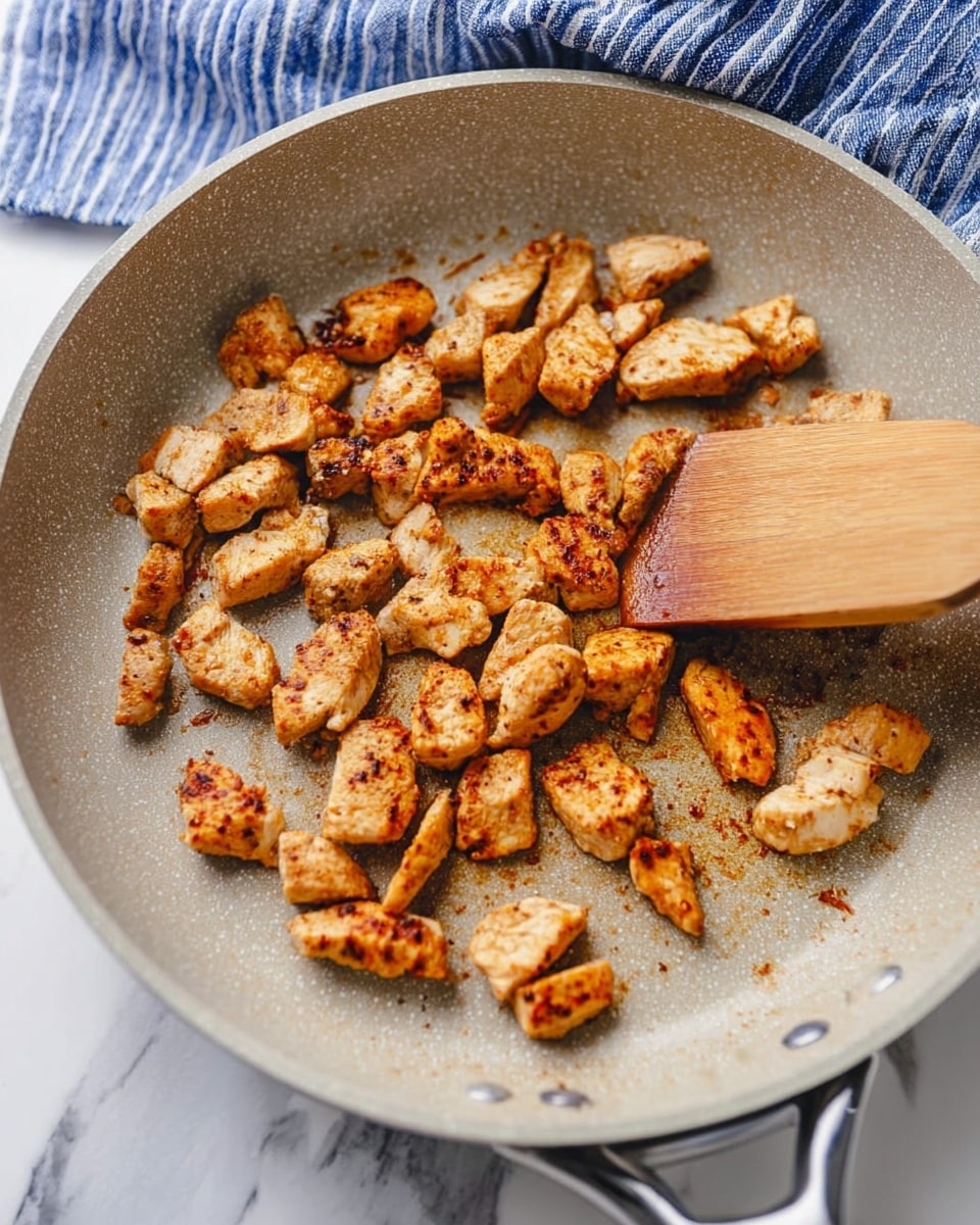 Small pieces of cooked chicken are scattered evenly across the bottom of a light grey non-stick pan. Each piece is golden brown with some darker spots, showing a crispy texture on the outside. On the right edge of the pan, a wooden spatula with a smooth finish is lifting some pieces. The pan is placed on a white marbled surface, and in the background, a blue and white striped cloth is slightly blurred. photo taken with an iphone --ar 4:5 --v 7