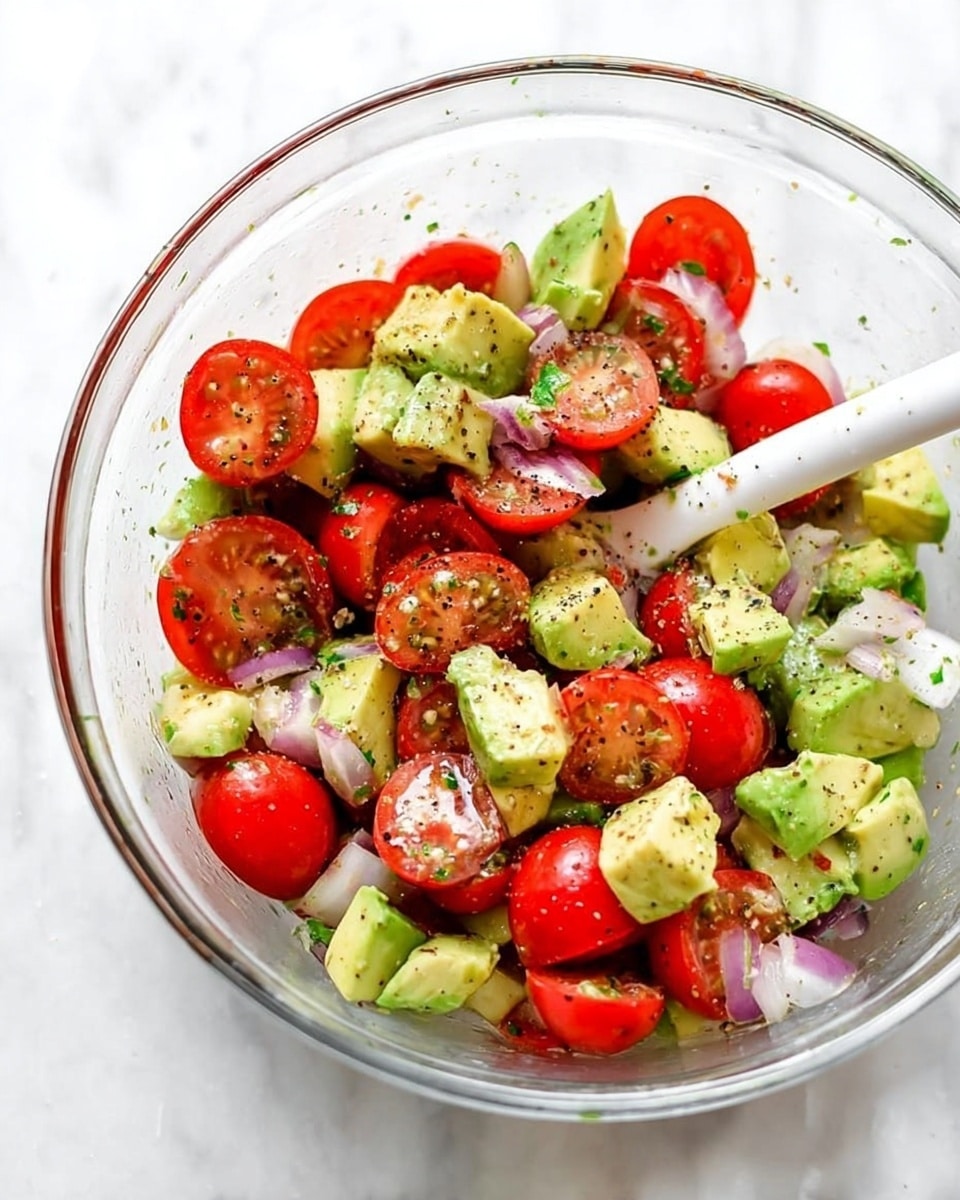 A clear glass bowl filled with a colorful salad composed of bright red halved cherry tomatoes, light green avocado chunks, and small pieces of pale purple onion mixed evenly throughout. The salad is lightly coated with a shiny dressing and sprinkled with coarse black pepper, giving the ingredients a fresh look. A white mixing spoon is visible inside the bowl resting among the ingredients. The bowl is set on a white marbled surface. Photo taken with an iphone --ar 4:5 --v 7