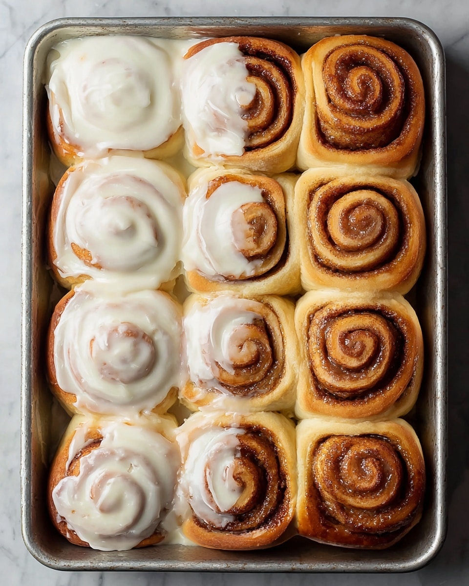 A metal baking tray holds eight cinnamon rolls arranged in two rows of four. The left half of the rolls is covered with a thick layer of creamy white icing that smoothly drapes over the warm golden-brown swirled dough, showing soft texture underneath. The right half of the rolls shows the golden to deep brown swirls of cinnamon sugar clearly visible on the baked dough, with a slightly crisp look around the edges, and a shiny glaze reflecting the light. The tray sits on a white marbled texture surface. photo taken with an iphone --ar 4:5 --v 7