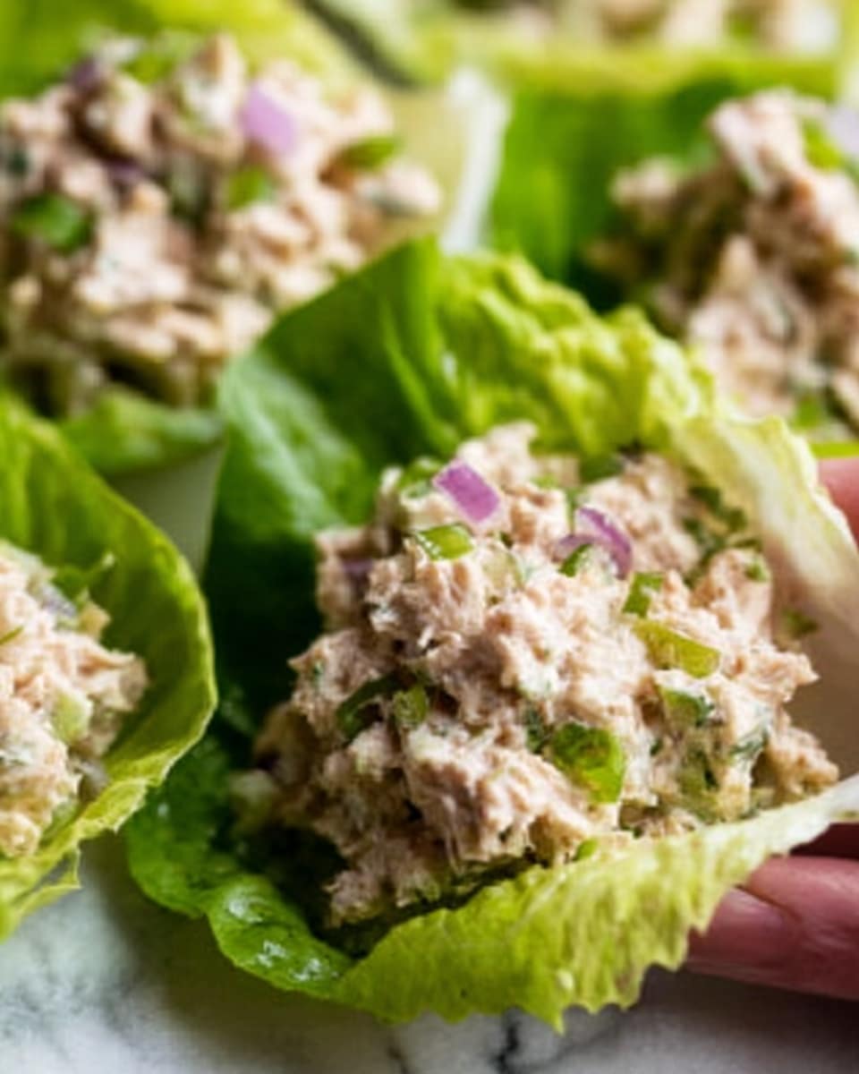 Three bright green lettuce leaves form a base on a large white plate lined with white parchment paper, each leaf holding a scoop of a pale, finely chopped mixture with small pieces of purple onion and bits of green herbs mixed in. The mixture has a light, creamy texture with specks of black pepper sprinkled on top. In the background, there are small white bowls with chopped ingredients and a bowl of reddish-brown sauce on a white marbled surface. Photo taken with an iphone --ar 4:5 --v 7