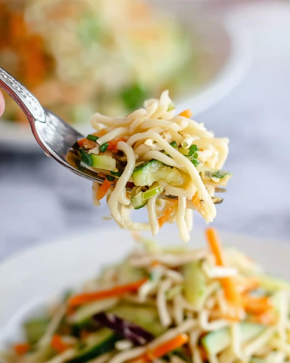 A close-up of a fork holding a small bite of noodle salad, with pale yellow noodles twisted and mixed with thin strips of light green cucumber, dark green scallion pieces, and thin orange carrot slices. Below the fork, a white plate filled with the same noodle salad is slightly blurred. The background has a soft white marbled texture, and a woman's hand is holding the fork at the left edge of the image. photo taken with an iphone --ar 4:5 --v 7