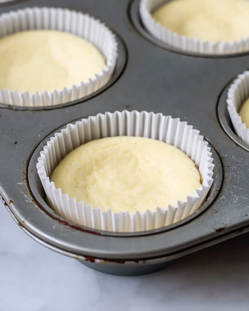 The image shows a close-up view of two cheesecake batter cups placed inside a gray muffin pan. Each cup has a white ridged paper liner filled with smooth, pale yellow cheesecake batter. The batter has a creamy texture with no toppings or decorations and sits evenly inside the paper liners. The pan rests on a white marbled surface, and there are faint marks and slight stains on the metal pan around the cups. The focus is on the front cup with the others softly blurred in the background. photo taken with an iphone --ar 4:5 --v 7