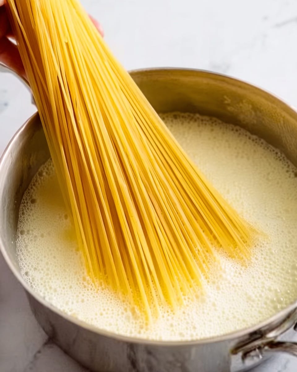 A silver pot filled with bubbling white liquid, likely boiling water or sauce, sitting on a white marbled surface. A large bunch of uncooked yellow spaghetti sticks is held by a woman's hand, being placed gently into the pot. The spaghetti strands are bright and straight, fanning out slightly as they are lowered into the creamy liquid. The scene focuses closely on the pot and the pasta, with no other objects around. Photo taken with an iphone --ar 4:5 --v 7