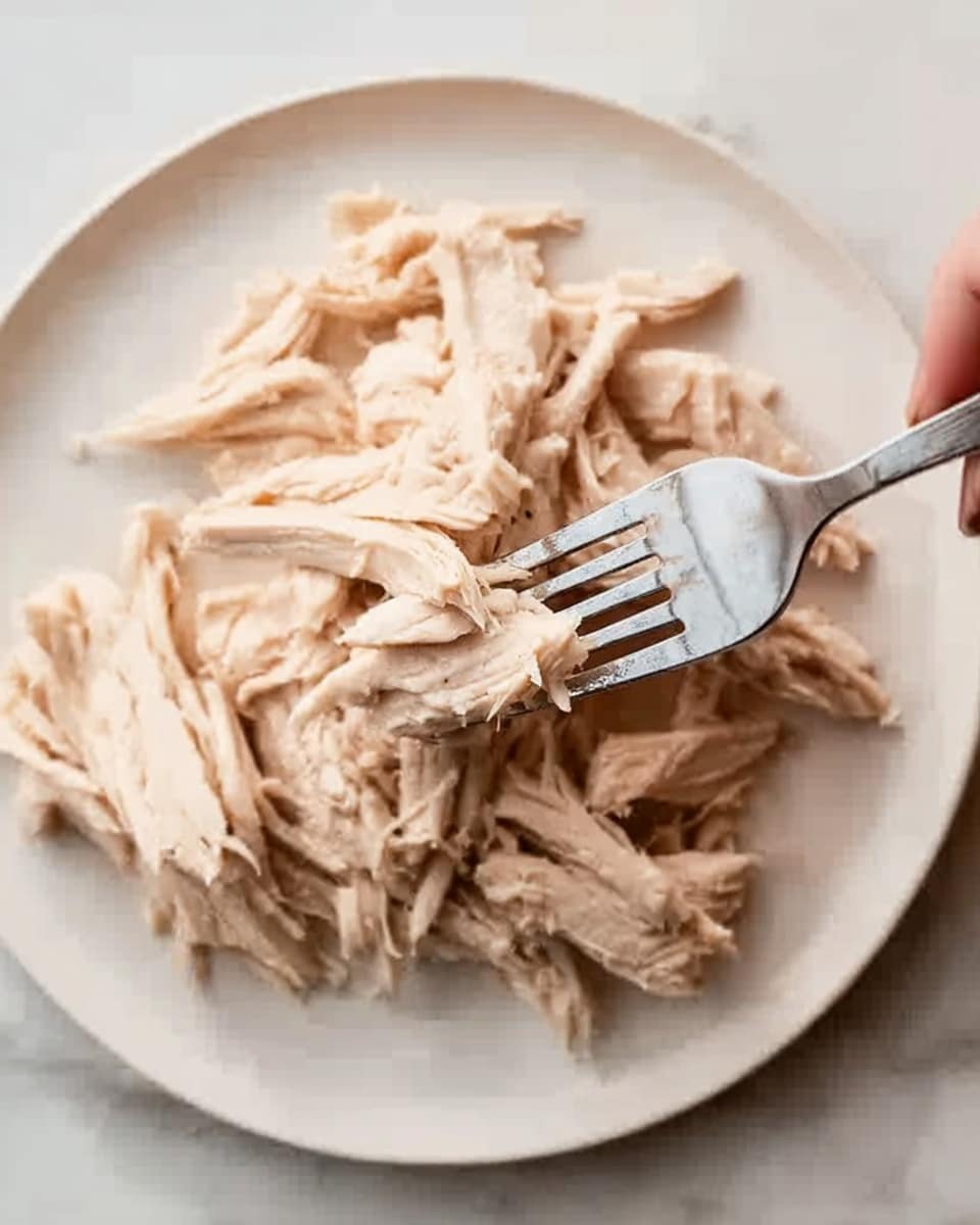 The image shows shredded cooked chicken on a white plate. The chicken is light beige in color with soft, fibrous texture, arranged in loose layers. A woman's hand holds a fork that is pulling apart the chicken, creating thin strips. The plate is on a white marbled surface, with bright natural light highlighting the tender meat. photo taken with an iphone --ar 4:5 --v 7