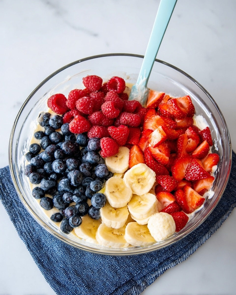 A clear glass bowl sits on a white marbled surface with a blue cloth underneath it. Inside the bowl, the bottom layer is a creamy white mixture partly visible around the edges. On top, there are four distinct groups of fresh fruit: bright red raspberries piled in one section, dark blue blueberries in another, yellow banana slices in the middle, and chopped red strawberries filling the remaining section. A light blue mixing spatula rests inside the bowl against the fruit. Photo taken with an iphone --ar 4:5 --v 7
