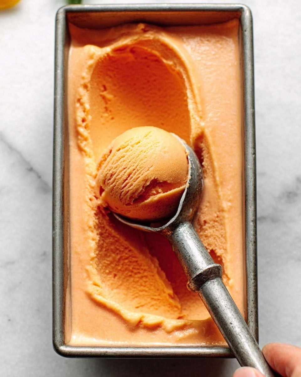 The image shows a rectangular metal container filled with smooth, orange-colored ice cream with a creamy texture. In the container, a vintage-style metal ice cream scoop holds a rounded scoop of the ice cream, showing slight ridges and a soft, thick texture. The background surface is white with a marbled texture, creating a clean and bright setting. A woman's hand can be seen gently holding the scoop's handle from the top right corner of the image. photo taken with an iphone --ar 4:5 --v 7