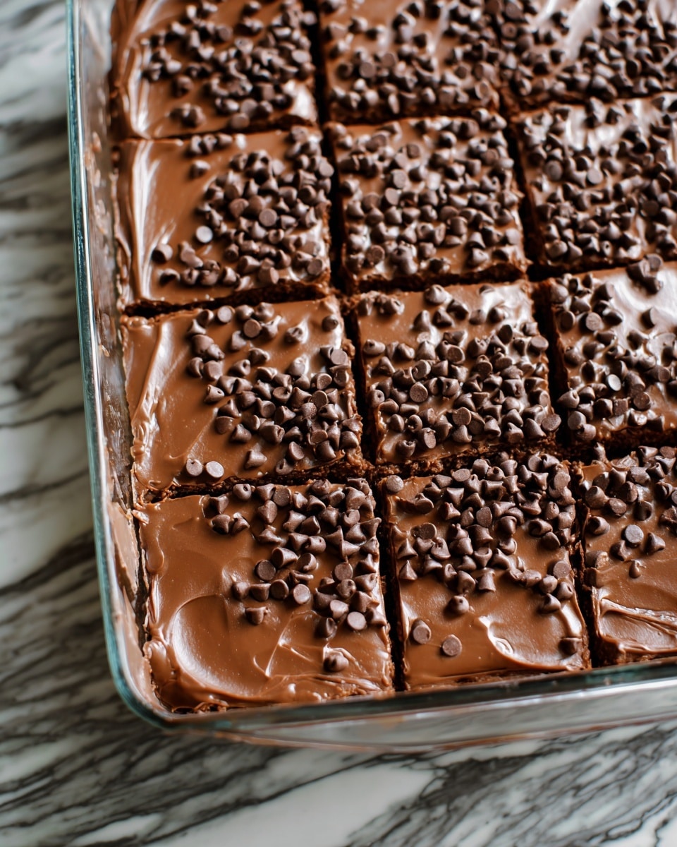 The image shows a glass baking dish filled with a rich, smooth layer of milk chocolate frosting topped thickly with small, shiny chocolate chips spread evenly across the surface. The dish is divided into eight rectangular pieces, with the lines of division faintly visible through the creamy frosting. The background surface is a white marbled texture that highlights the glossy chocolate layers. photo taken with an iphone --ar 4:5 --v 7