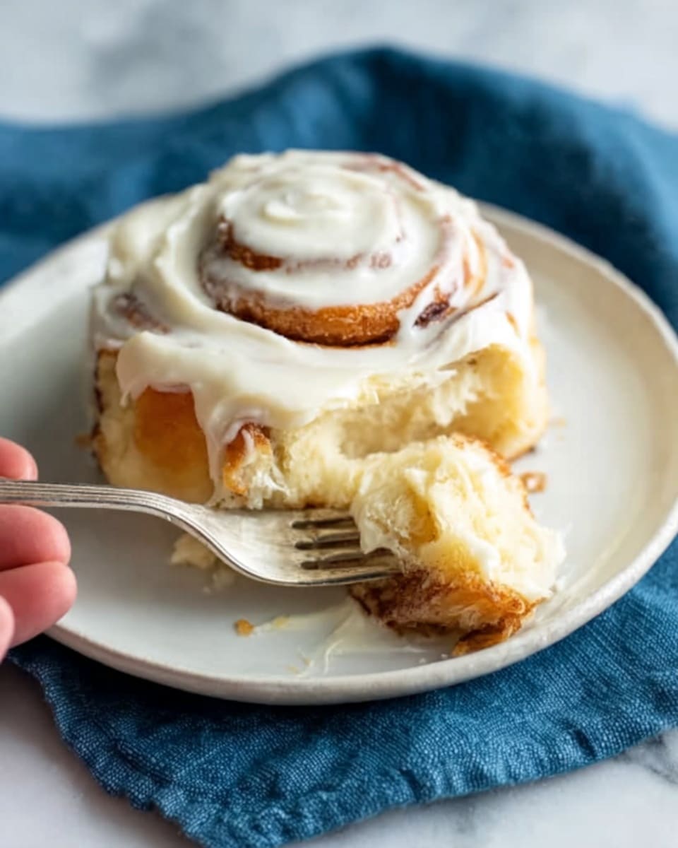 A soft cinnamon roll sits on a white plate over a white marbled surface, topped with thick creamy white icing that spreads over the sides. The roll is golden brown with swirls of cinnamon visible beneath the icing. A woman's hand holds a piece of the roll on a silver fork, showing the fluffy inside texture. A blue cloth is partially visible under the plate, adding a simple contrast. photo taken with an iphone --ar 4:5 --v 7