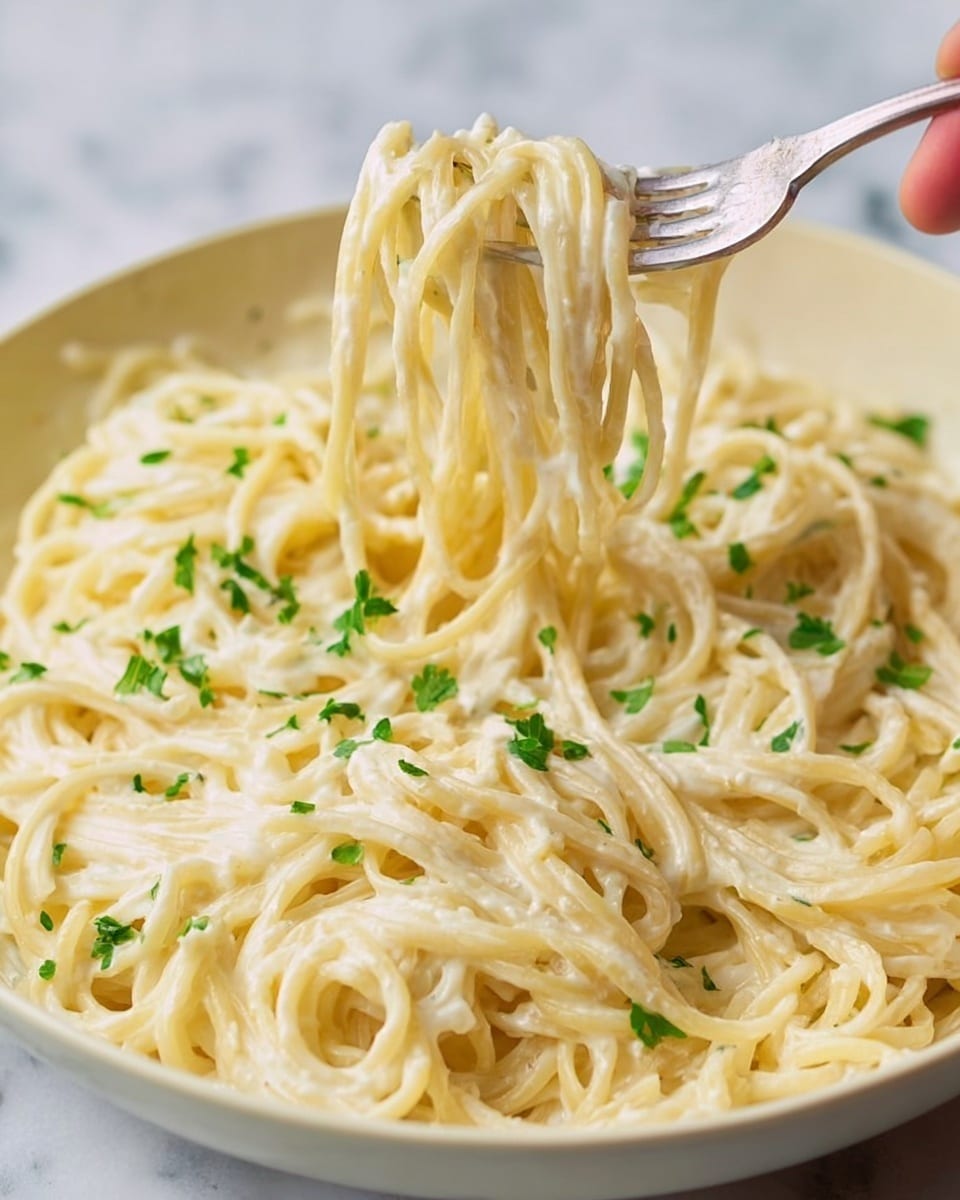 The image shows a close-up of creamy spaghetti pasta in a white bowl on a white marbled surface. The spaghetti strands are coated evenly with a thick, smooth white sauce. Small pieces of fresh green herbs are sprinkled on top, adding a touch of color. A woman's hand is holding a fork that is twirling some of the spaghetti, lifting it slightly above the bowl with visible texture in the sauce and pasta strands. The background remains soft and blurred to keep the focus on the pasta. photo taken with an iphone --ar 4:5 --v 7